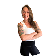 Smiling woman with arms crossed, wearing a white tank top and black shorts; colorful tattoos on both arms visible.