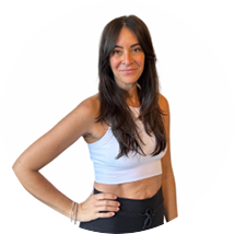 Smiling woman with long dark hair in a light blue crop top and dark pants, standing with one hand on her hip.