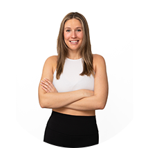 Woman standing against a white background, smiling with arms crossed, wearing a white sleeveless top and black pants.