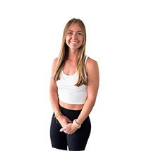 Smiling woman with long blonde hair wearing a white crop top and black pants, standing with hands clasped in front.