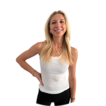Smiling woman with long blonde hair wearing a white tank top and black pants, standing with one hand on her hip against a white background.
