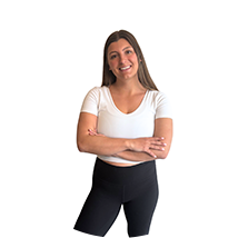 A woman standing with arms crossed, smiling, wearing a white t-shirt and black shorts.